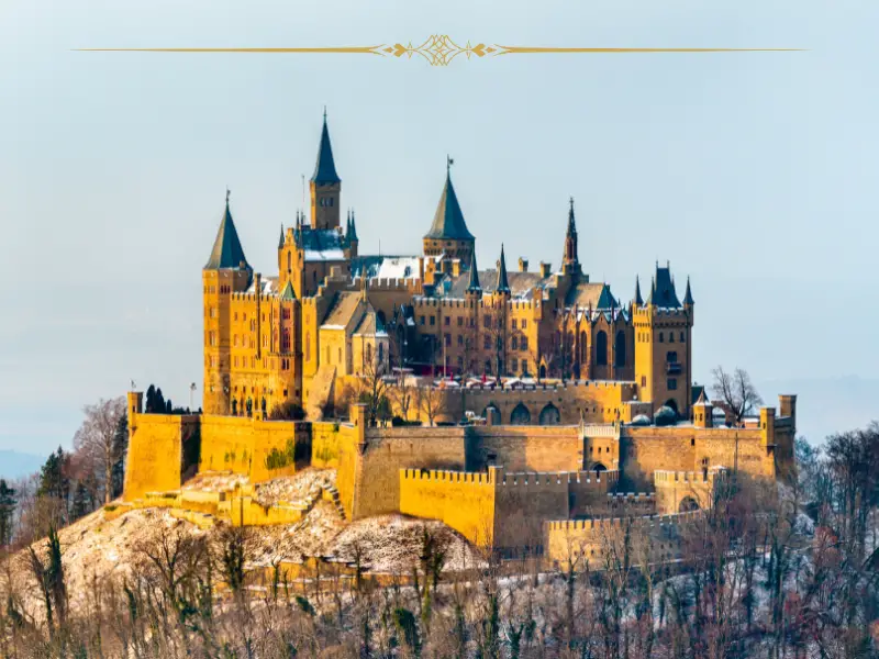 An impressive large castle on top of a hill. It has a lot of towers and a defense wall around it. The photo has been taken in winter, at sunset. The light is very warm on the castle, making it look yellow. There is snow on the ground.