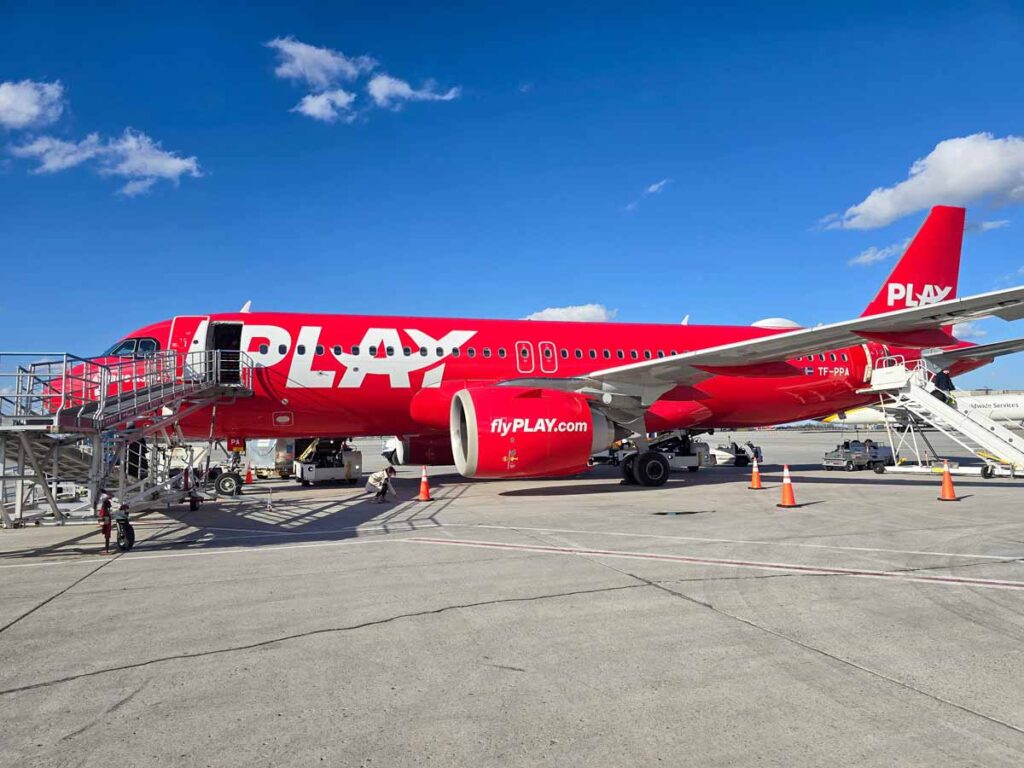 A red plane with Play written in white on it, on the tarmac at Hamilton airport. The sky is blue, it's a very sunny day.