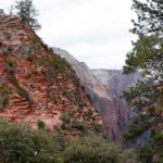 A view in Zion National Park. On the left there is a large rock, almost orange, with people climbing it. On the right of the rock there is a big drop. The weather is cloudy.