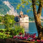 A castle on the shore of a lake seen from the shore in front, through trees, and flowers. Behind the castle there is the slop of a mountain, covered with green trees.
