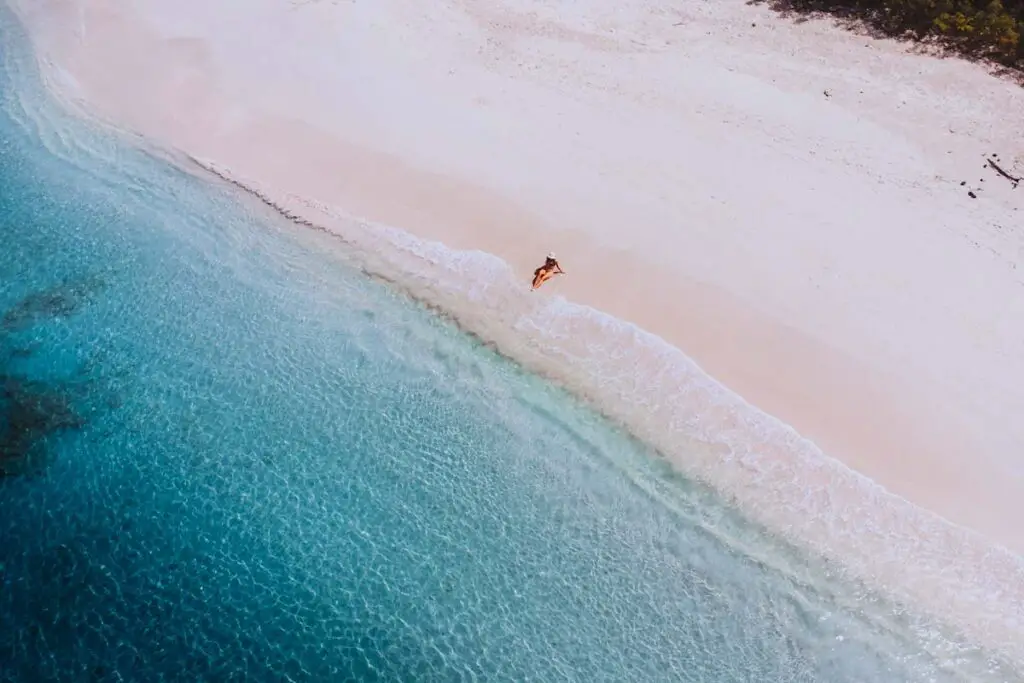A drone photo of a large sandy beach with blue, clear water. There is only one woman on the beach, sitting down on the shore, with her feet in the water. She is wearing a red bathing suit and a white had