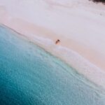 A drone photo of a large sandy beach with blue, clear water. There is only one woman on the beach, sitting down on the shore, with her feet in the water. She is wearing a red bathing suit and a white had