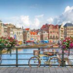 A view of a canal in Amsterdam, with a bike tied to the railings of a bridge. Behind you can see houses with colourful facades.