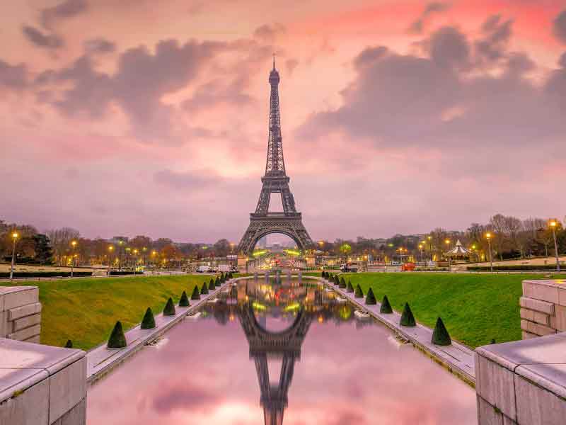 Eiffel Tower with a fountain of water in front of it, in which it reflects and a pink sky. The photo has been taken at sunrise and there are no people around.