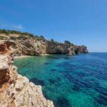 The sea next to the cliffs in Ibiza. The sea has several shades of blue. The sky is blue, without any clouds. The rocks are bare and quite tall