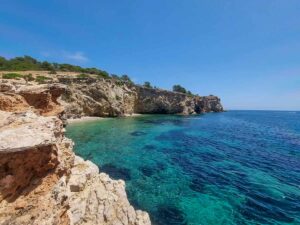 The sea next to the cliffs in Ibiza. The sea has several shades of blue. The sky is blue, without any clouds. The rocks are bare and quite tall