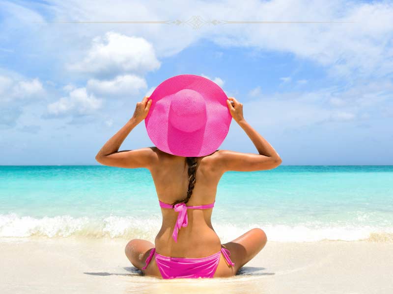A woman wearing a pink bathing suit and a pink hat, holding the hat with her hands, sitting on the beach with the back at the camera. In front of her there is the ocean.