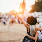 Two girls hugging from the side, rising their plastic cups filled with beer. In front of them, blurred, you can see a crowd of people and the sun setting, making an orange background.