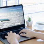a person using a laptop trying to book a flight. On the screen there is a plane and a booking flight form. Next to the laptop there is a cup of coffee and a small plant.