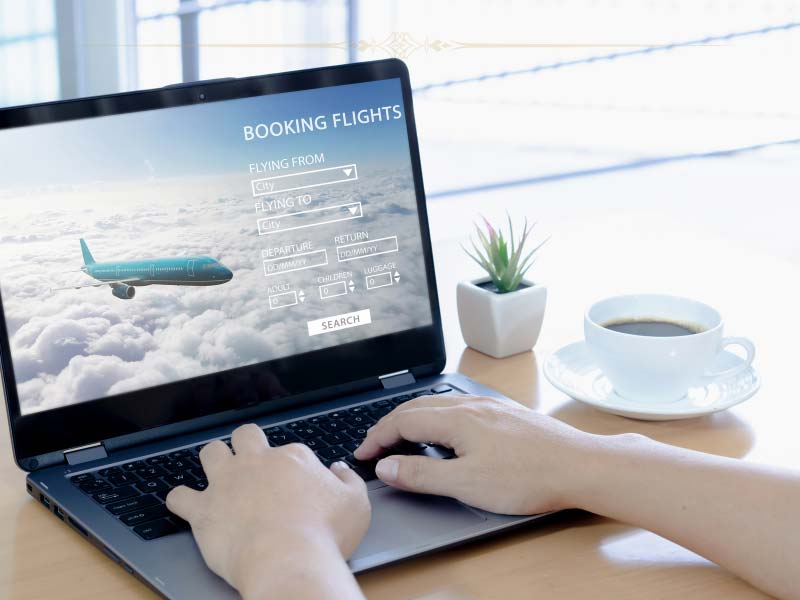 a person using a laptop trying to book a flight. On the screen there is a plane and a booking flight form. Next to the laptop there is a cup of coffee and a small plant.
