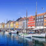 Colorful buildings alongside a canal, where white boats and anchored. It is a beautiful sunny day. The boats reflect in the water underneath them.