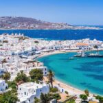 A white town on a Greek island, inside a bay. There are a few boats and a larger ship anchored by the pier.
