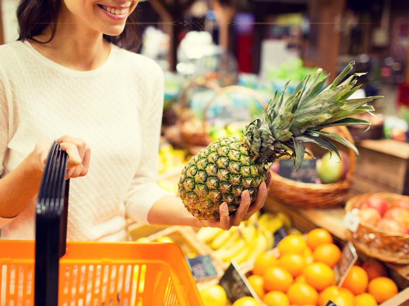 A woman holding a large pineapple in one hand and an orange shopping basket in the other.