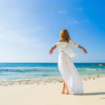 What Is The New Quiet Travel Trend And Why Is It Becoming So Popular? A woman dressed in a white dress standing on a beautiful sandy beach. She has her arms spread in the wind.