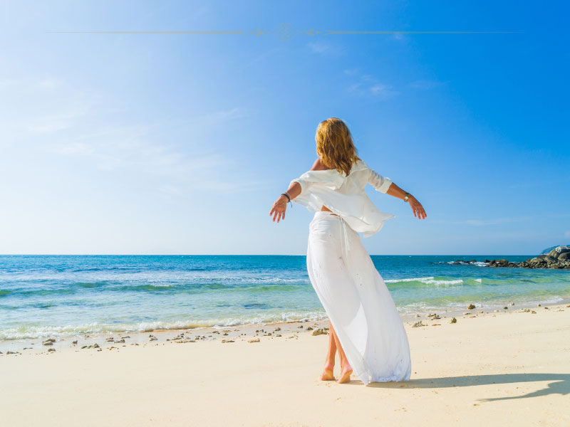 A woman dressed in a white dress standing on a beautiful sandy beach. She has her arms spread in the wind.