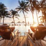 Two people sitting in beach chairs with the back at the camera, in front of a pool, looking at the sunset through the palms in front of them.