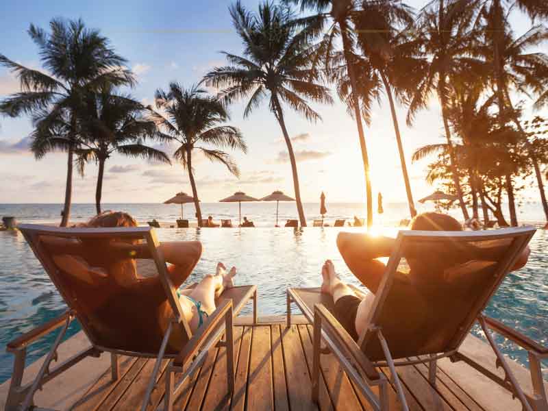 Two people sitting in beach chairs with the back at the camera, in front of a pool, looking at the sunset through the palms in front of them.