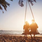 Passport to Longevity: How Travel Can Add Years to Your Life A couple sitting together on a swing on the beach, with their backs at the camera, watching the sunset. The sun is in front of them, making them look like silhouettes.