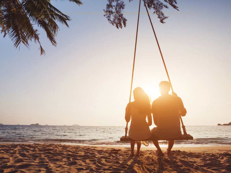 A couple sitting together on a swing on the beach, with their backs at the camera, watching the sunset. The sun is in front of them, making them look like silhouettes.