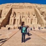 I am posing in front of the main temple at Abu Simbel. I am wearing a green tshirt, black jeans, and a black jacket. I have a camera around my neck. You can see four large statues behind me, with the entrance to the temple underneath