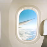 A close-up of a window on the plane. You can see the wing and clouds below.