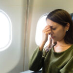A woman sitting next to a window, on a plane, wearing a green jumper, looking visibly worried.
