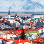 A very small city located on the shores of the water. Behind you can see a mountain covered with snow. The houses are small, and they have red rooftops.
