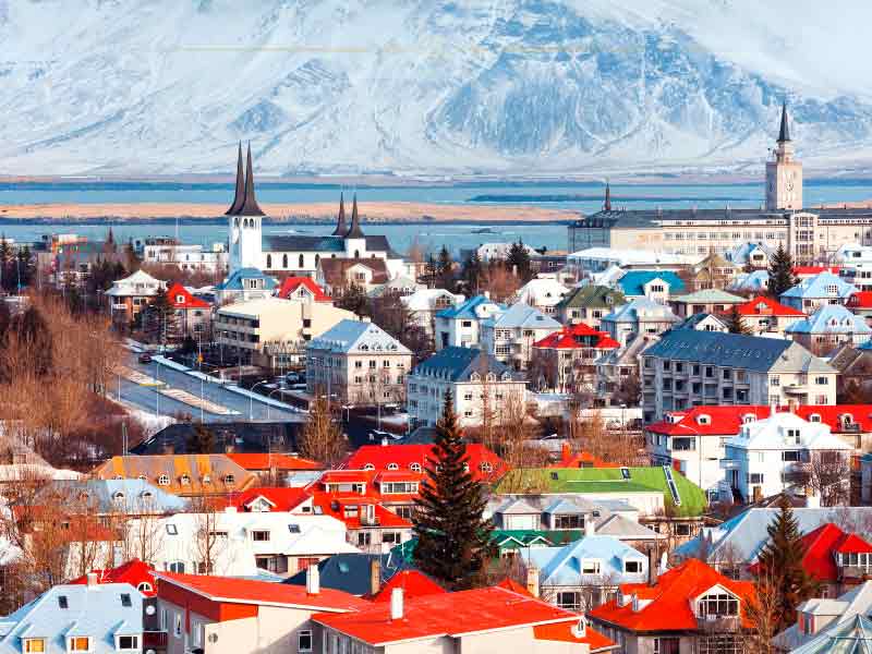 A very small city located on the shores of the water. Behind you can see a mountain covered with snow. The houses are small, and they have red rooftops.