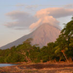 A large volcano seen from a beach lined with palm trees, in Nicaragua. It's sunset time and the atmosphere is very orange.