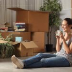 A woman sitting on the floor with a cup of coffee in her hands. She is wearing blue jeans and a grey top. Around her there are packing boxes and a few plants.