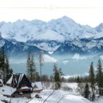 Two houses with triangle shaped rooftops, covered with snow. In the background you can see tall mountains covered with snow.