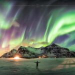 A man looking at the northern light, with his open arms. In front there are green and pink shades on the sky, above a mountain.