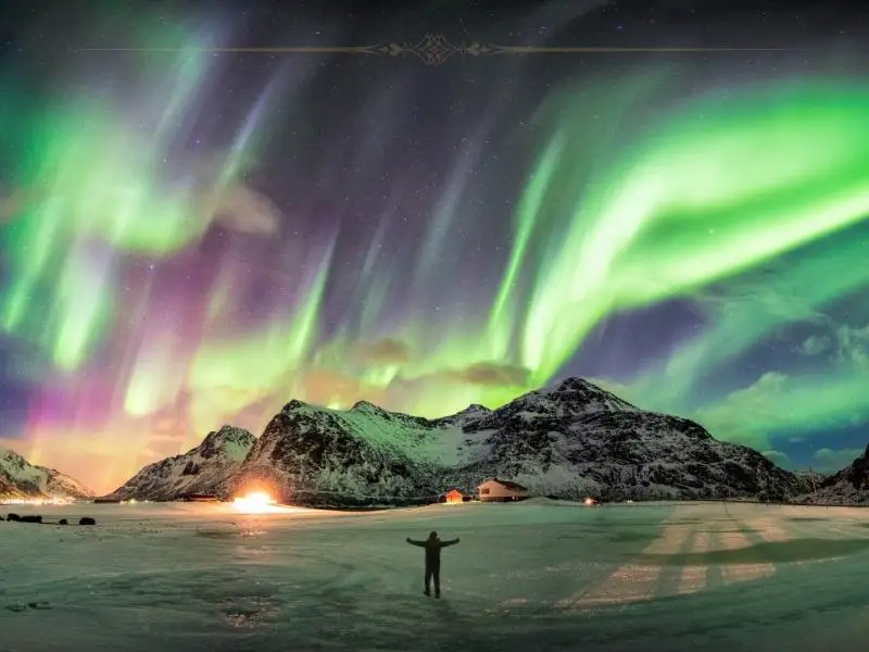 A man looking at the northern light, with his open arms. In front there are green and pink shades on the sky, above a mountain.