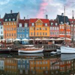 Colourful houses reflecting in the water next to them. There are small boats anchored by the shore.