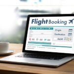 A close-up of a laptop with a flight booking engine opened on it. Next to the laptop there is a white cup.