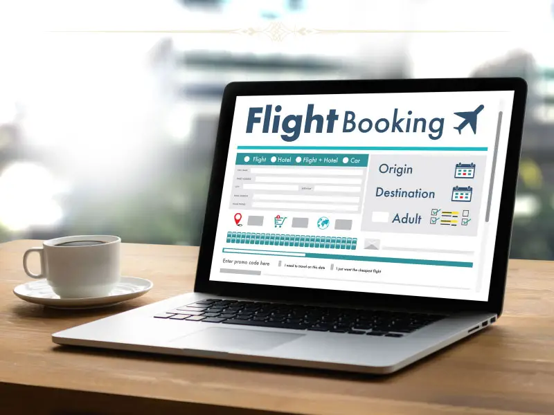 A close-up of a laptop with a flight booking engine opened on it. Next to the laptop there is a white cup.
