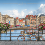 A photo of Amsterdam on a sunny day, with typical houses along the canal, and a bike tied to the side of a bridge.