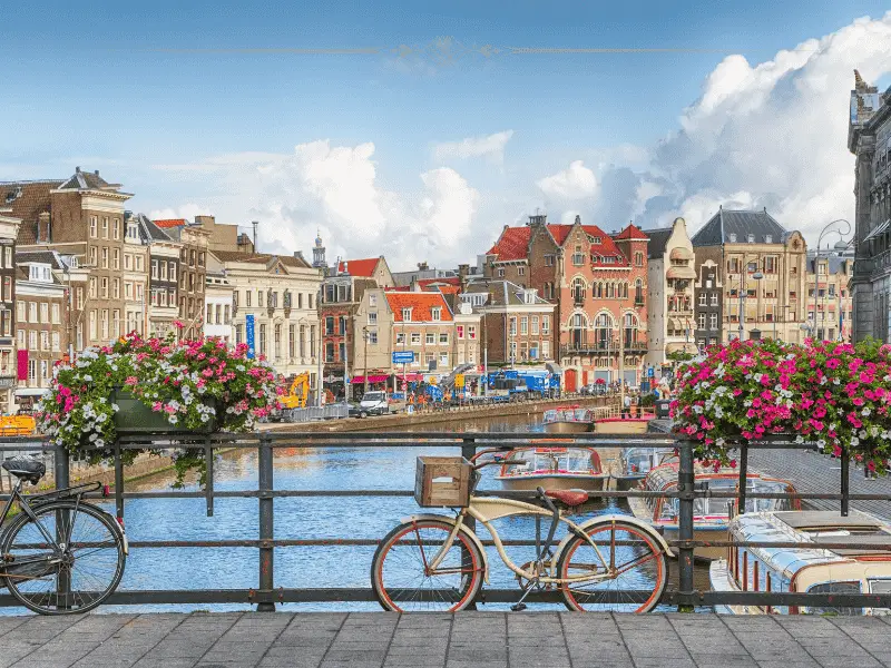 A photo of Amsterdam on a sunny day, with typical houses along the canal, and a bike tied to the side of a bridge.
