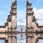 A woman standing between two structures, with her hands up in the air. She is wearing a red dress. The image is reflected below in what looks like water.