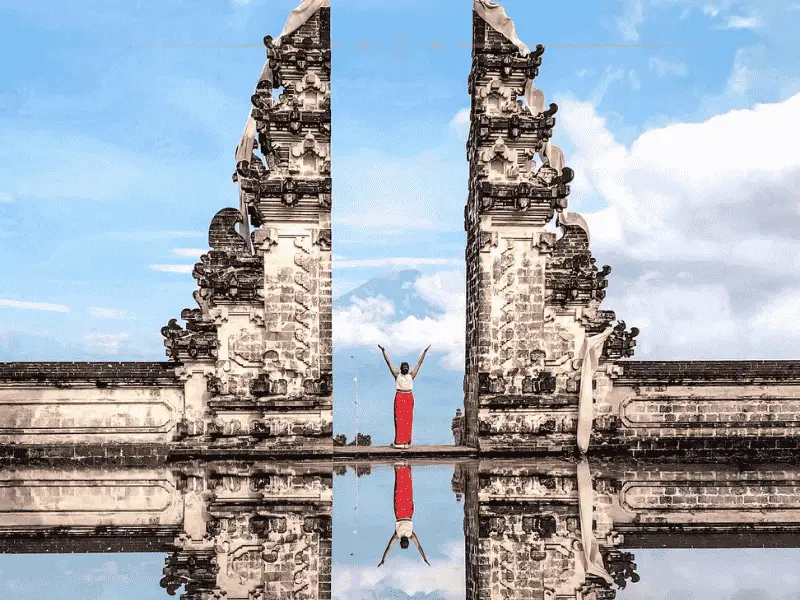 A woman standing between two structures, with her hands up in the air. She is wearing a red dress. The image is reflected below in what looks like water.