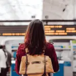 A woman wearing a dark red blouse and a light brown backpack, with the back at the camera, looking at a departure board.