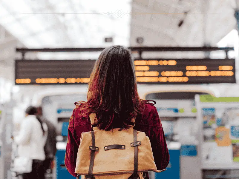 A woman wearing a dark red blouse and a light brown backpack, with the back at the camera, looking at a departure board.