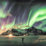 A man holding his hands open for the Northern lights in front of him which light the sky green.