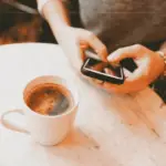 A close-up of a woman texting in a cafe, with a cup of coffee on the table next to her.