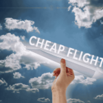 A close-up of a hand holding a paper plane with cheap flights written on it, on a background of a sky with fluffy clouds on it.