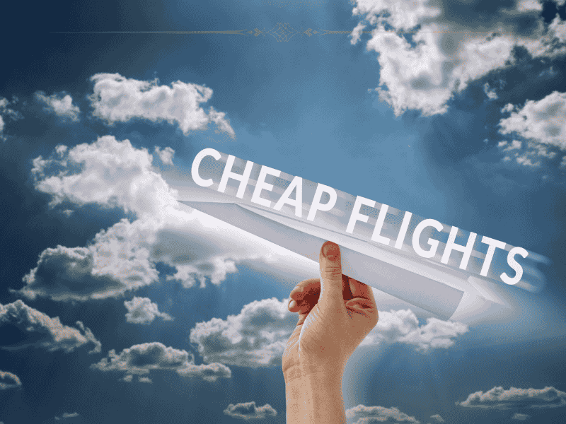 A close-up of a hand holding a paper plane with cheap flights written on it, on a background of a sky with fluffy clouds on it.