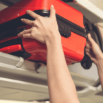 A close-up of a pair of hands pushing a red bag inside the overhead compartment on a plane.