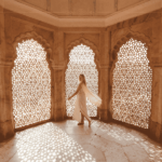 A woman wearing a white flowy dress inside an Indian palace, with typical windows covered by sculpted wood.