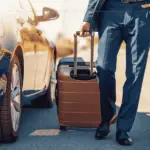 A close-up of the bottom part of a man wearing a suit, dragging a smart suitcase behind him. He is walking next to a car.