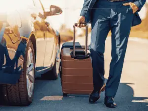 A close-up of the bottom part of a man wearing a suit, dragging a smart suitcase behind him. He is walking next to a car.